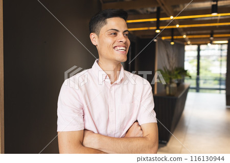 A biracial young man wearing a pink shirt is standing, arms crossed, laughing 116130944