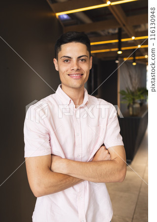 A young biracial man standing with arms crossed, wearing pink shirt in a modern business office A young biracial man standing with arms crossed, wearing pink shirt in a modern business office 116130948