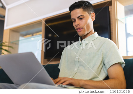 Biracial young man working on laptop, sitting on couch in a modern business office Biracial young man working on laptop, sitting on couch in a modern business office 116131036