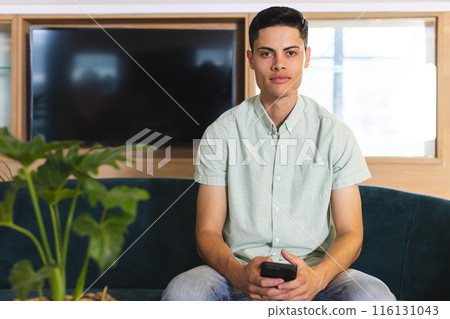 A young biracial man holding a smartphone sits on a couch in a modern business office 116131043