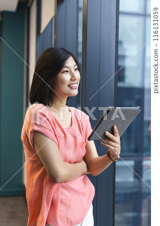 A young Asian woman holding a tablet and looking away in a modern business office 116131059