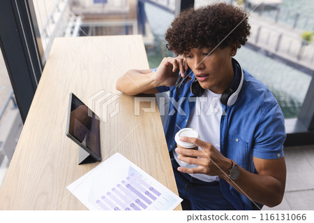A young biracial man with curly brown hair is talking on the phone in a modern business office 116131066
