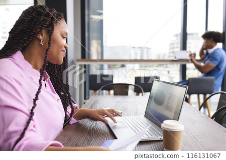 A young biracial woman with braided hair is working on a laptop in a modern business office 116131067