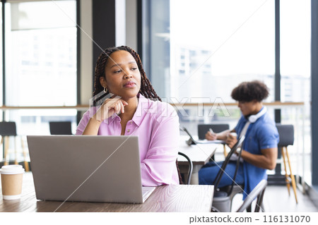 A young biracial woman with braided hair is working on a laptop in a modern business office 116131070