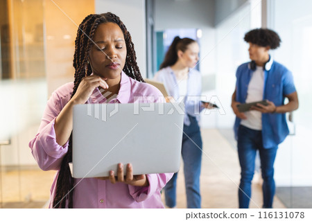 Biracial woman holding laptop, thinking, colleagues walking behind in a modern business office 116131078