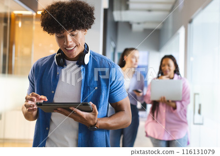 Biracial young man holding tablet in a modern business office, two women behind using tech 116131079