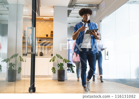 Young biracial man walking, using tablet and drinking coffee in a modern business office 116131082