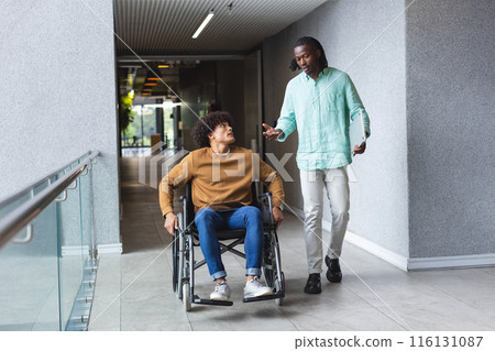 African American man pushing wheelchair of biracial man, both talking in a modern business office 116131087