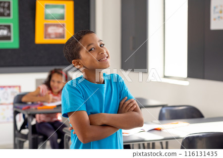 In school, young biracial boy standing with arms crossed, looking thoughtful in the classroom In school, young biracial boy standing with arms crossed, looking thoughtful in the classroom 116131435