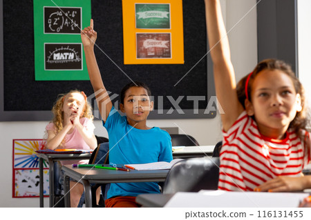In school, diverse group of young students sitting at desks in a classroom, raising hands 116131455