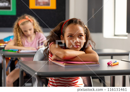 In school, young biracial female student sitting at a desk in a classroom, looking thoughtful 116131461