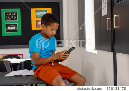 In school, in a classroom, a young biracial boy is sitting, focusing on a smartphone In school, in a classroom, a young biracial boy is sitting, focusing on a smartphone 116131478
