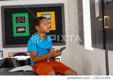 In school, in classroom, young biracial boy holding a smartphone, looking at it In school, in classroom, young biracial boy holding a smartphone, looking at it 116131484