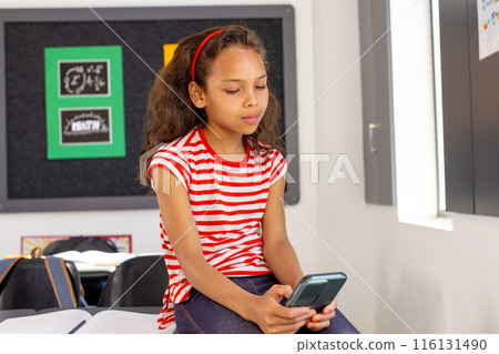In school, young biracial girl holding a smartphone sits at a desk in a classroom 116131490