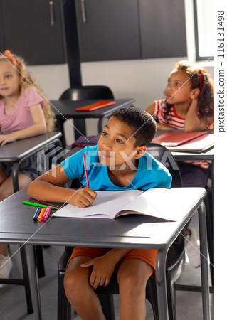 In school, young biracial male student sitting at a desk in a classroom, writing 116131498