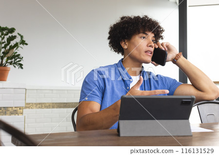 Young biracial man talking on phone, looking away, sitting at table in a modern business office Young biracial man talking on phone, looking away, sitting at table in a modern business office 116131529