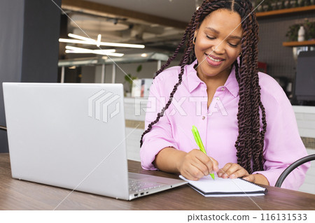 A young biracial woman wearing a pink shirt is writing in a notebook in a modern business office 116131533