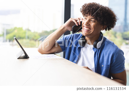 Biracial young man talking on phone, wearing headphones around neck in a modern business office 116131534