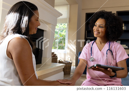 Mature biracial woman holding tablet, talking to young nurse at home 116131603