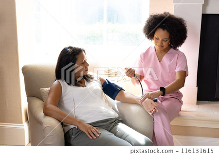 Mature biracial woman getting blood pressure checked by young biracial nurse at home 116131615