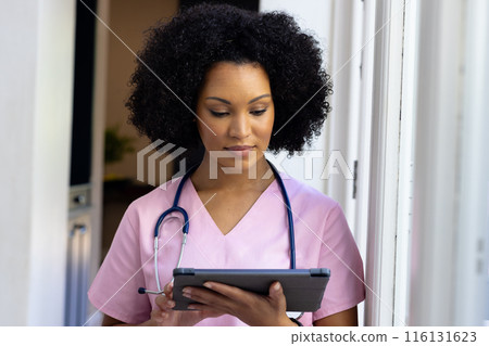 Young biracial nurse wearing pink scrubs checking tablet at home Young biracial nurse wearing pink scrubs checking tablet at home 116131623