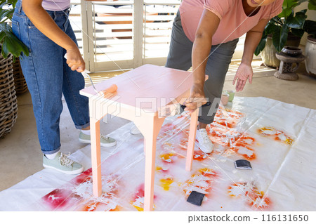 Mature biracial woman painting stool at home in an upcycling project, young biracial woman assisting 116131650