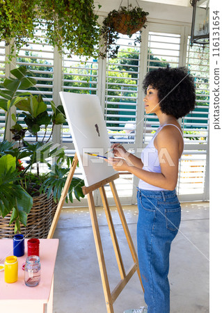 Biracial young woman painting on canvas at home, surrounded by plants 116131654