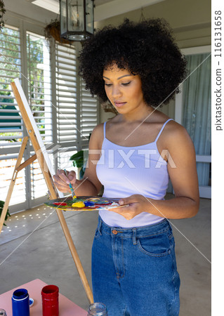 Biracial young woman painting on canvas at home, mixing colors on palette 116131658