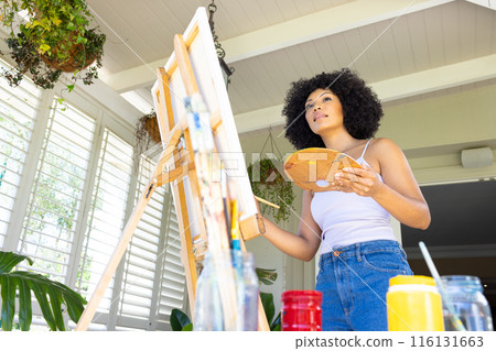 Young biracial woman painting on canvas at home, holding palette Young biracial woman painting on canvas at home, holding palette 116131663