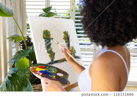 Biracial young woman painting on canvas at home, focusing intently 116131666