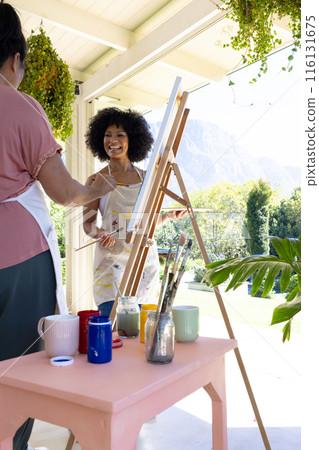 Mature biracial woman painting at home, young biracial woman watching, both smiling 116131675