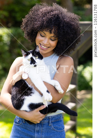 Biracial young woman holding black and white cat, both outdoors at home in garden 116131708
