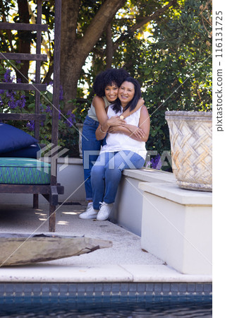 Biracial mother and adult daughter are embracing on a garden bench at home in the garden 116131725