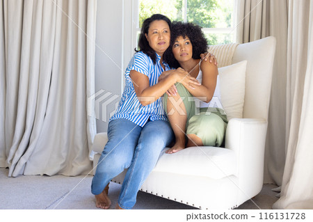 Mature biracial woman hugging young adult daughter, both sitting at home 116131728