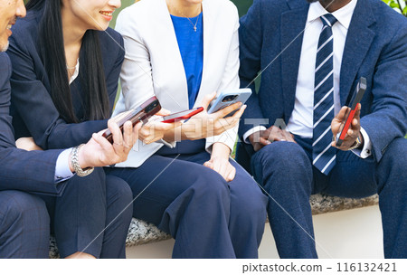 A group of multinational business people sitting on an outdoor bench and looking at their smartphones 116132421