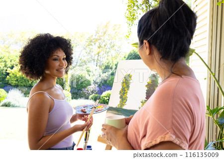 Mother and Daughter, mature biracial woman holding coffee mug watching young biracial woman painting 116135009