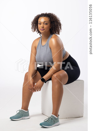 Biracial young female plus size model wearing sportswear sitting on white block, on white background Biracial young female plus size model wearing sportswear sitting on white block, on white background 116135056
