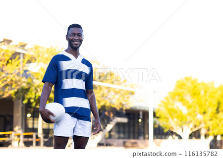 African American young male athlete holding a rugby ball, standing on field outdoors, copy space 116135782