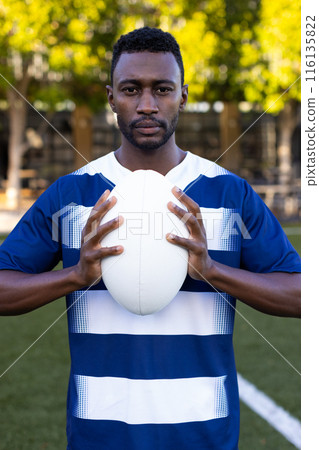 African American young male athlete holding a rugby ball on field outdoors, looking at camera African American young male athlete holding a rugby ball on field outdoors, looking at camera 116135822