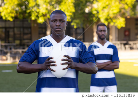 Two African American young male athletes are holding a rugby ball on a field outdoors 116135839