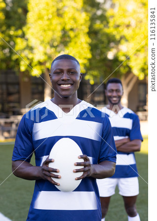 Two African American young male athletes are holding a rugby ball, smiling, on a field outdoors 116135841