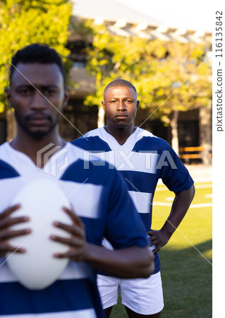 Two African American young male athletes are holding a rugby ball on field outdoors, looking focused 116135842
