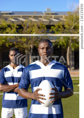Two African American young male athletes, holding a rugby ball, standing on field outdoors 116135850