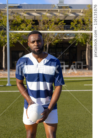 African American young male athlete holding rugby ball on field outdoors, looking serious African American young male athlete holding rugby ball on field outdoors, looking serious 116135876