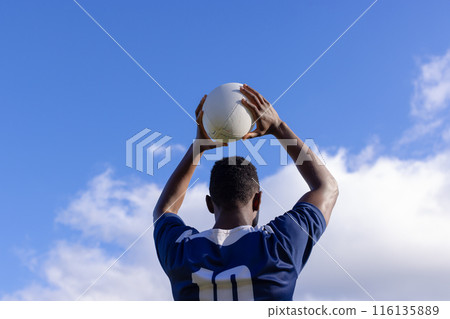 African American young male athlete holding white rugby ball above head on field, copy space 116135889
