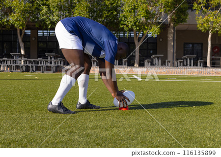 African American young male athlete placing rugby ball on grass on field, wearing sportswear 116135899
