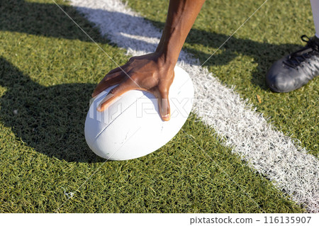 An African American young male athlete wearing cleats placing hand on rugby ball on grass field 116135907
