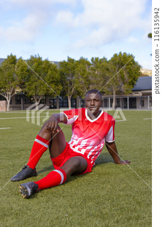 An African American young male athlete sitting on grass on field outdoors, wearing soccer gear 116135942