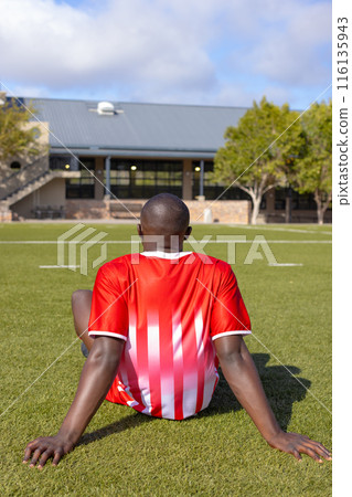 African American young male athlete sitting on grass on field outdoors, facing away African American young male athlete sitting on grass on field outdoors, facing away 116135943