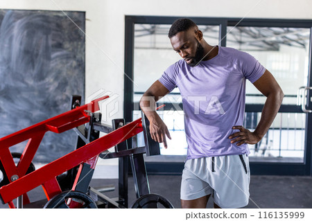African American young male athlete standing next to gym equipment in gym, looking thoughtful African American young male athlete standing next to gym equipment in gym, looking thoughtful 116135999